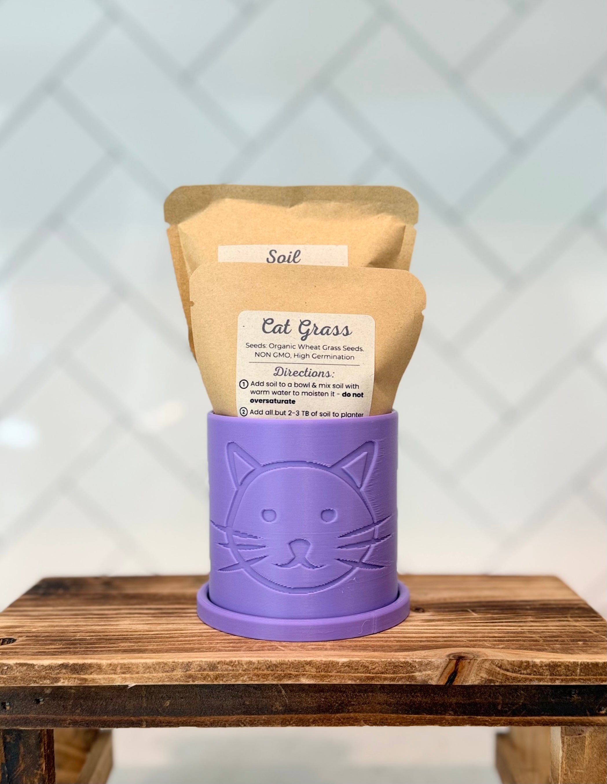 Bright Lilac colored planter with cat head design containing packaged cat grass seeds and soil on a wooden surface.