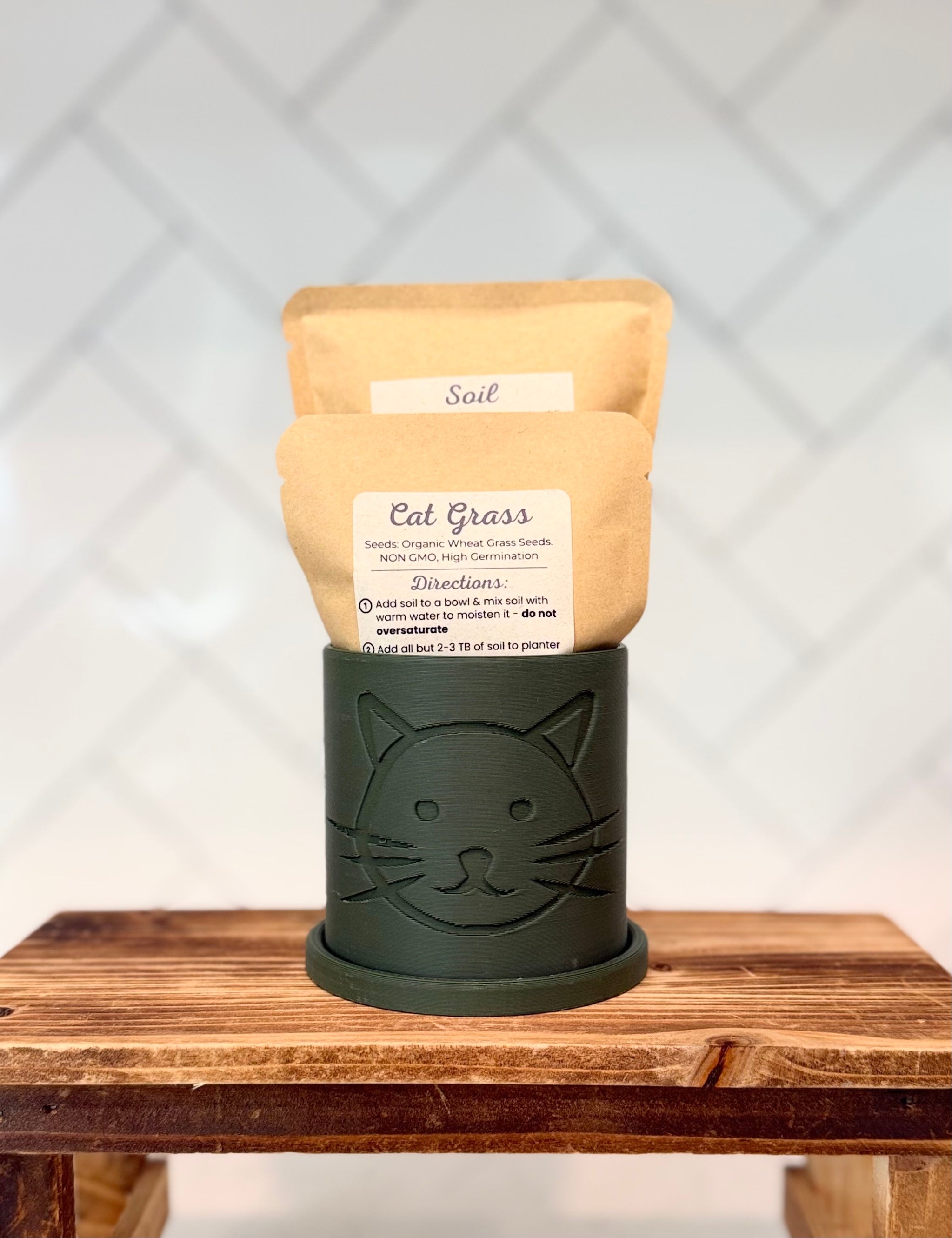 Dark Green colored planter with cat head design containing packaged cat grass seeds and soil on a wooden surface.