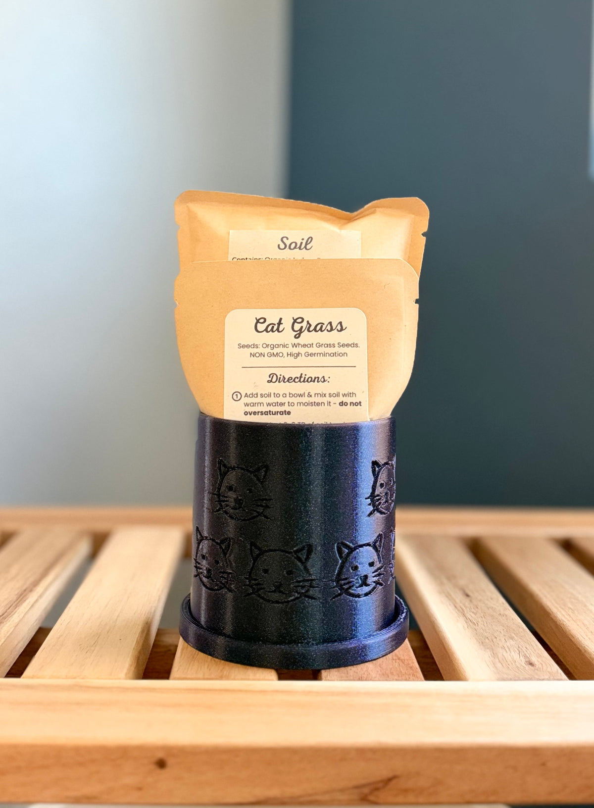 A small shimmering midnight blue colored cylindrical planter with a cat face pattern sits on a wooden slatted surface against a gray-blue background. Inside the planter are two pouches - a medium-sized packet labeled 'Cat Grass Seeds', and a larger packet labeled 'Soil.