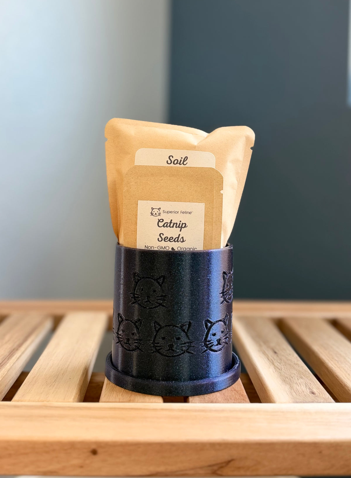 A small shimmering deep midnight blue colored cylindrical planter with a cat face pattern sits on a wooden slatted surface against a gray-blue background. Inside the planter are two pouches - a small packet labeled 'Catnip Seeds', and a larger packet labeled 'Soil.