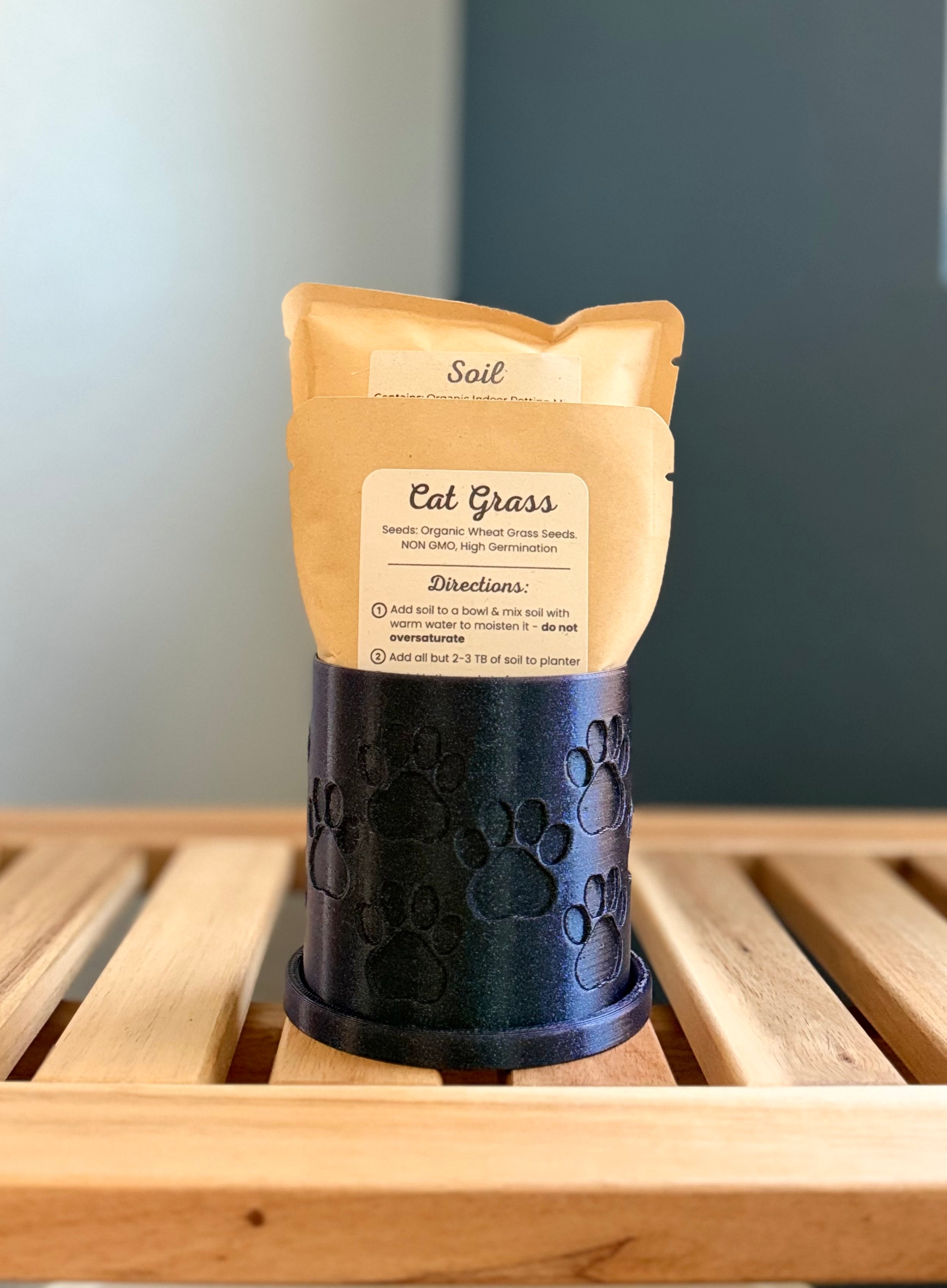 A small shimmering midnight blue colored cylindrical planter with a paw print pattern sits on a wooden slatted surface against a gray-blue background. Inside the planter are two pouches - a medium-sized packet labeled 'Cat Grass Seeds', and a larger packet labeled 'Soil.