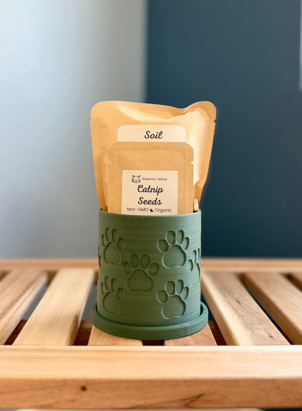 A green cylindrical planter with embossed paw prints sits on a wooden slatted surface. Inside the planter are two pouches - a small packet labeled 'Catnip Seeds', and a larger packet labeled 'Soil.'