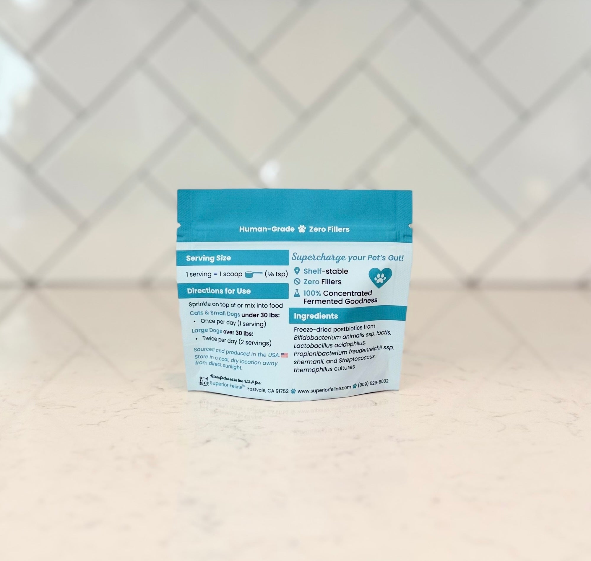 Back side of Superior Feline Microbiome Booster packaging with text about directions for use, ingredients and serving size on a kitchen countertop with a tiled backsplash