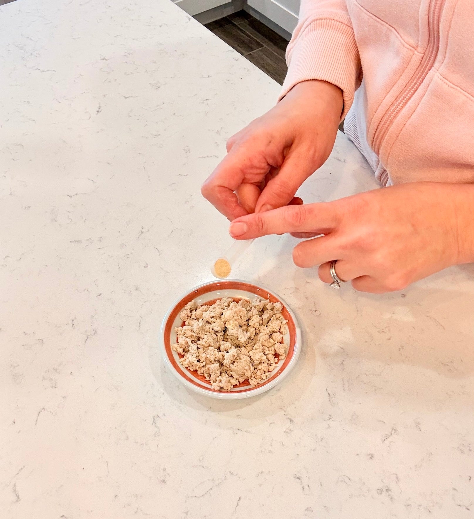 Person sprinkling Microbiome Booster over a small bowl of pet food on a light-colored surface