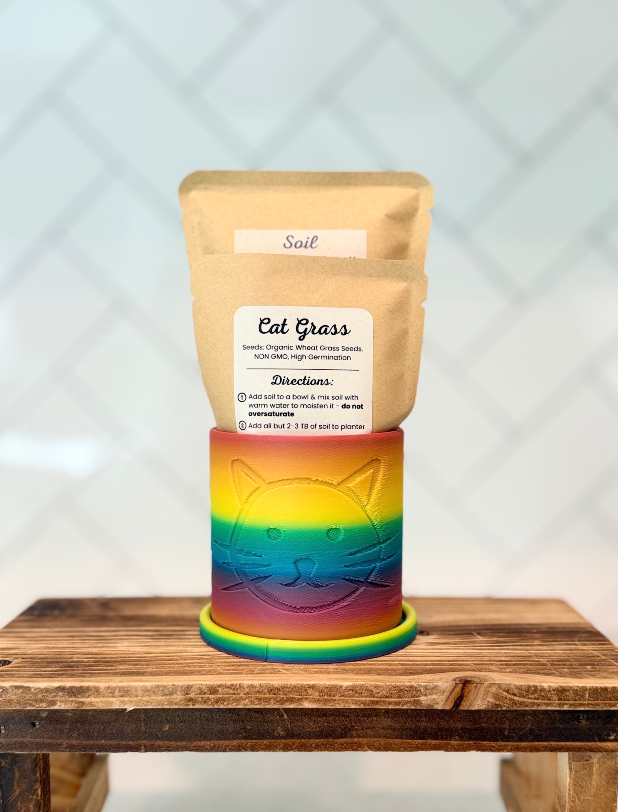 Rainbow colored planter with cat head design containing packaged cat grass seeds and soil on a wooden surface.
