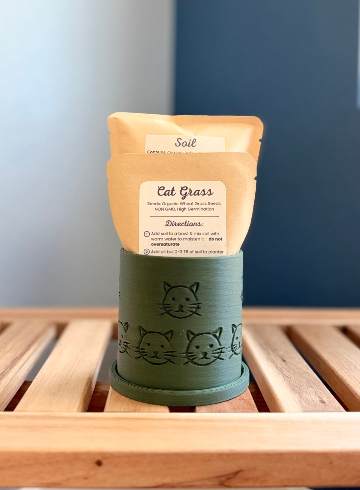 A small green colored cylindrical planter with a cat face pattern sits on a wooden slatted surface against a gray-blue background. Inside the planter are two pouches - a medium-sized packet labeled 'Cat Grass Seeds', and a larger packet labeled 'Soil.