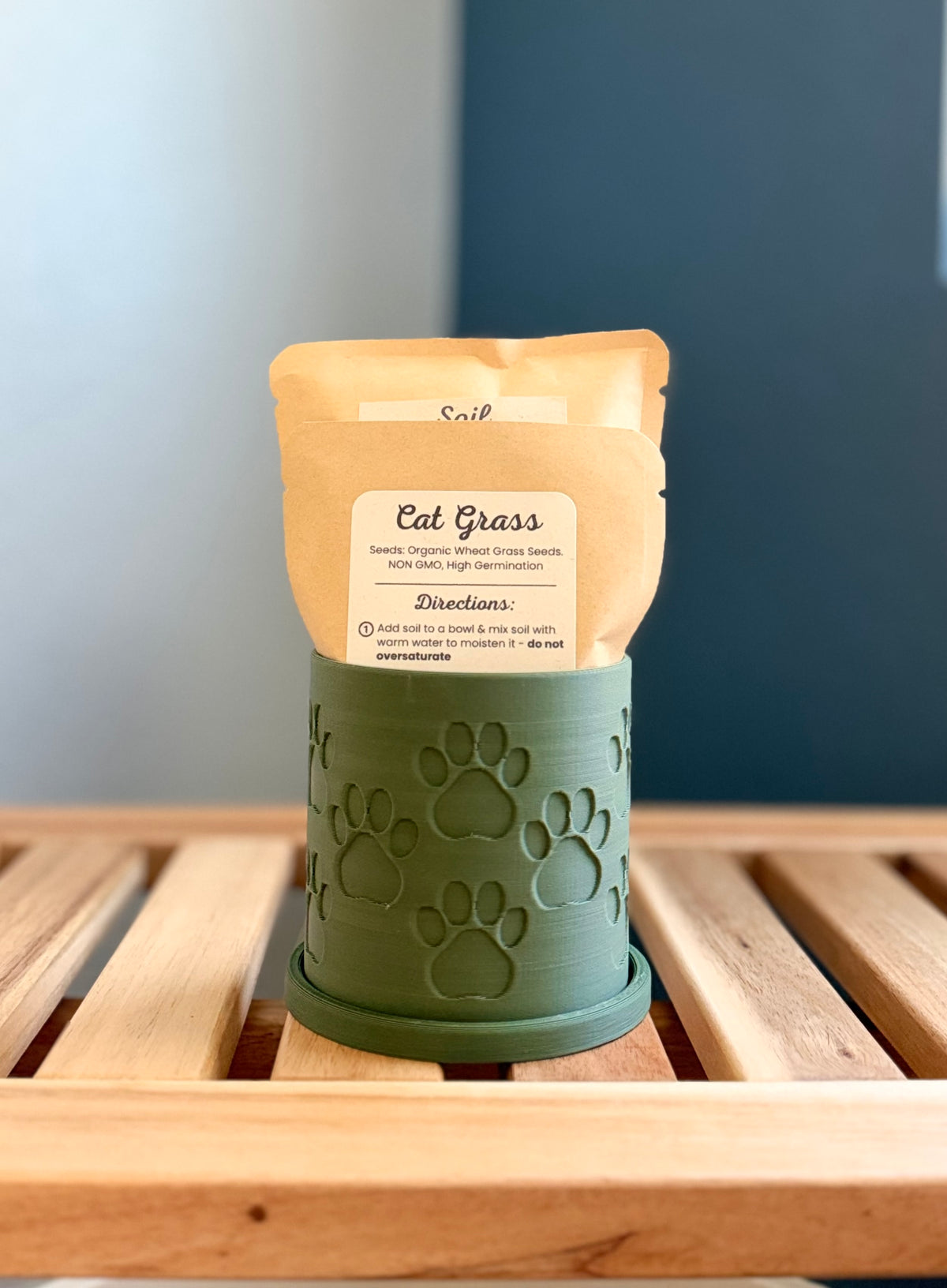 A small green colored cylindrical planter with a paw print pattern sits on a wooden slatted surface against a gray-blue background. Inside the planter are two pouches - a medium-sized packet labeled 'Cat Grass Seeds', and a larger packet labeled 'Soil.