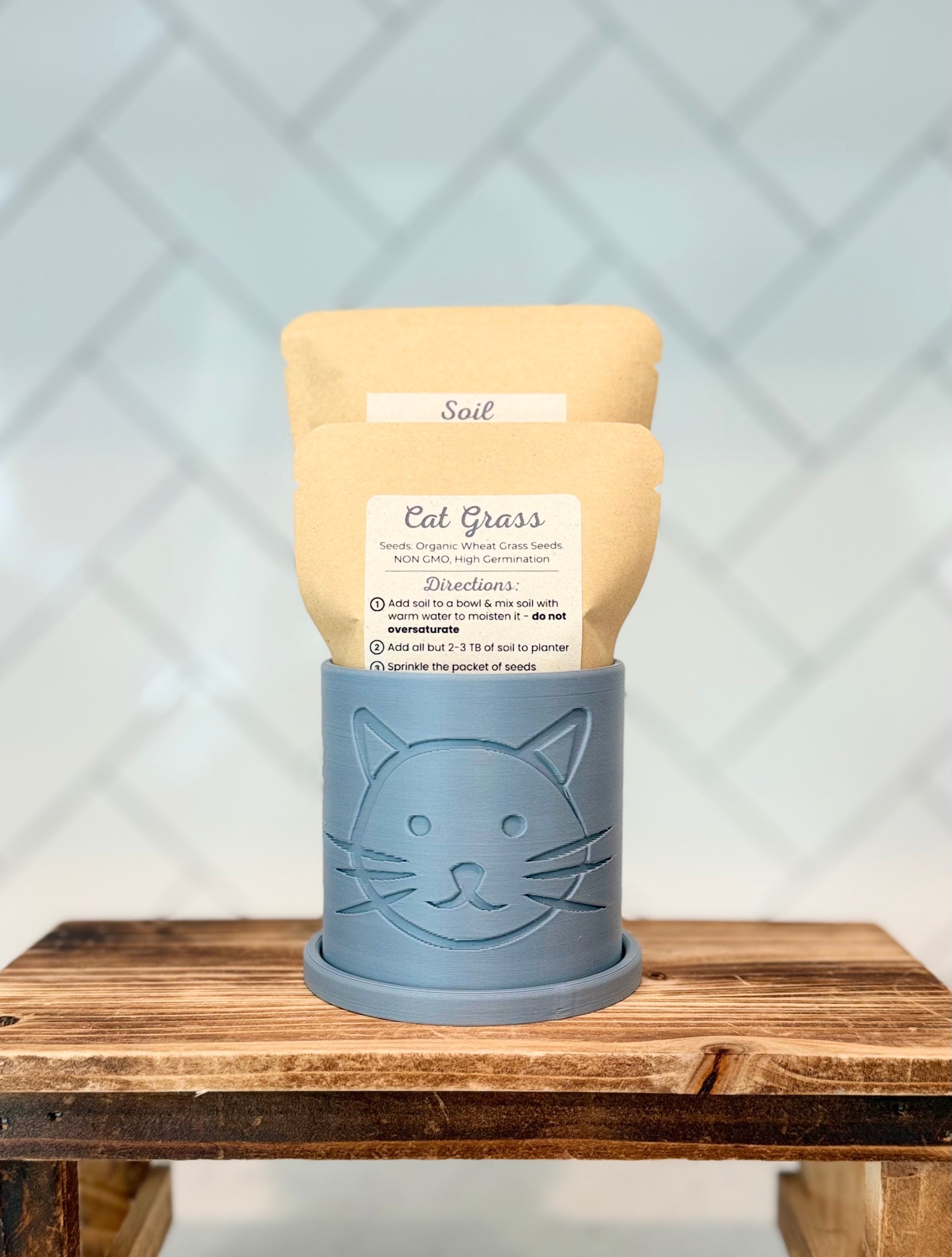 Gray colored planter with cat head design containing packaged cat grass seeds and soil on a wooden surface.