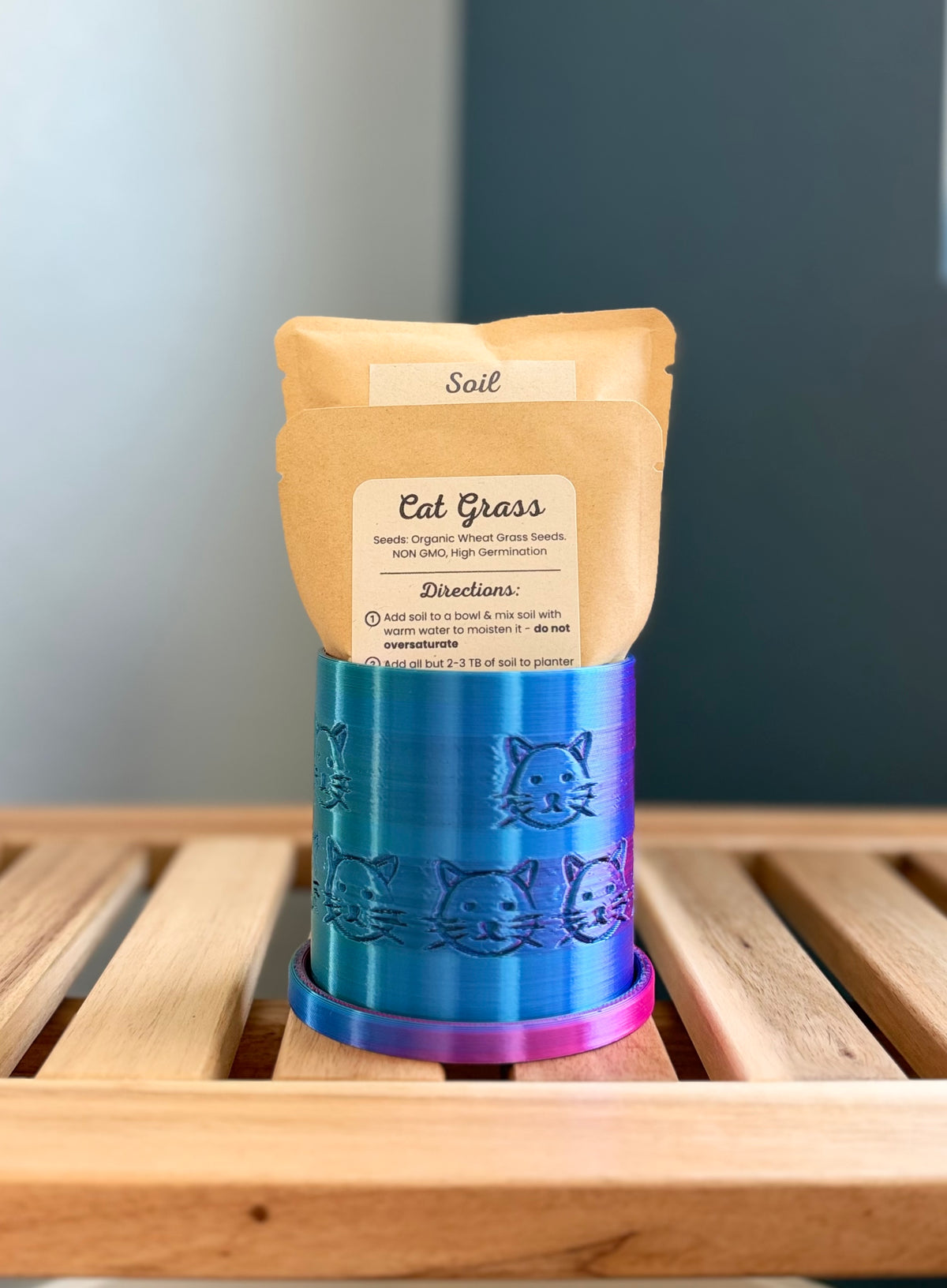 A small metallic iridescent colored cylindrical planter with a cat face pattern sits on a wooden slatted surface against a gray-blue background. Inside the planter are two pouches - a medium-sized packet labeled 'Cat Grass Seeds', and a larger packet labeled 'Soil.