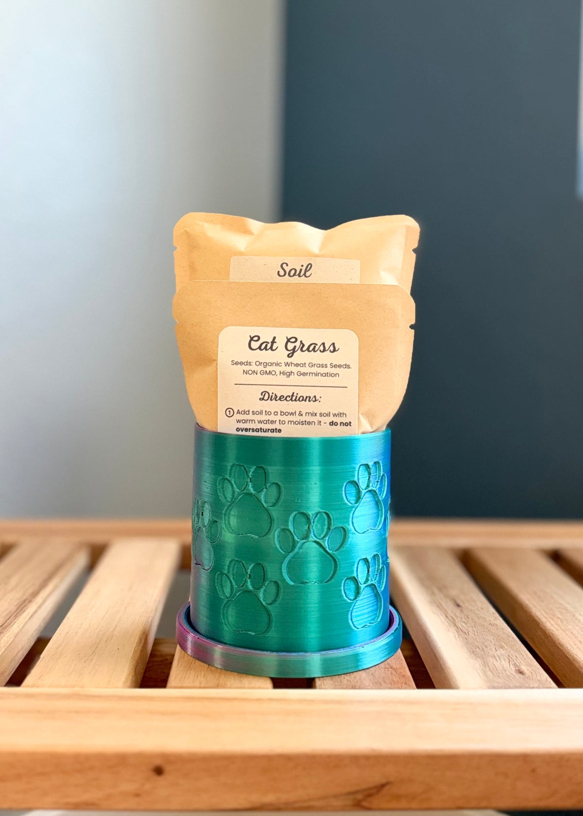 A small metallic iridescent colored cylindrical planter with a paw print pattern sits on a wooden slatted surface against a gray-blue background. Inside the planter are two pouches - a medium-sized packet labeled 'Cat Grass Seeds', and a larger packet labeled 'Soil.