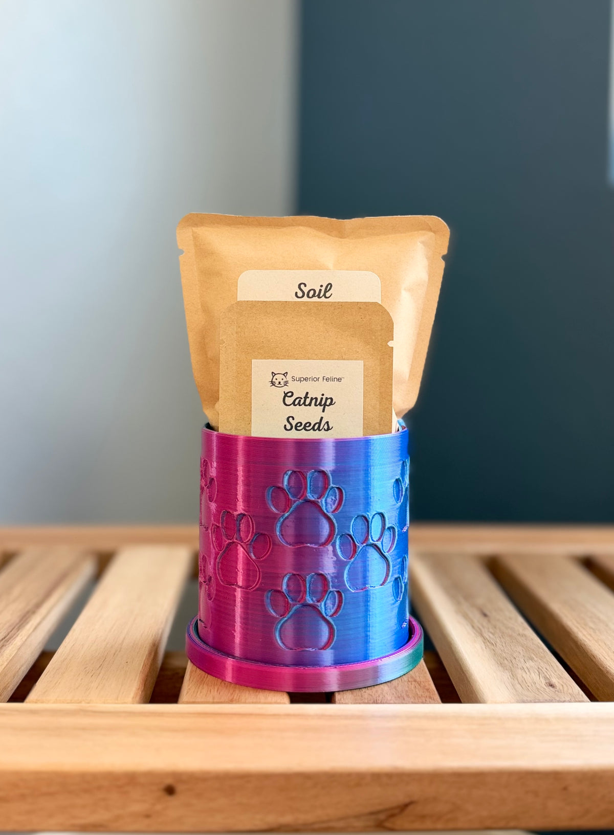 A small biodegradeable 3D-printed PLA planter with a pink-to-blue iridescent finish and embossed paw prints, holding two kraft paper packets labeled Soil and Catnip Seeds, displayed on a wooden slatted surface.